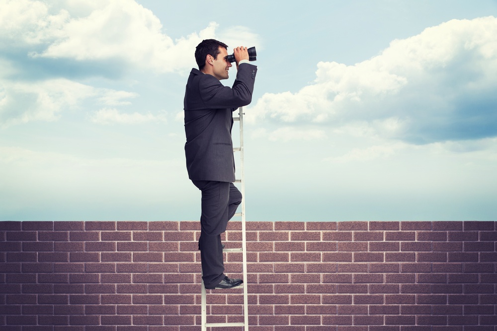 Businessman standing on ladder against blue sky over a brick wall.jpeg