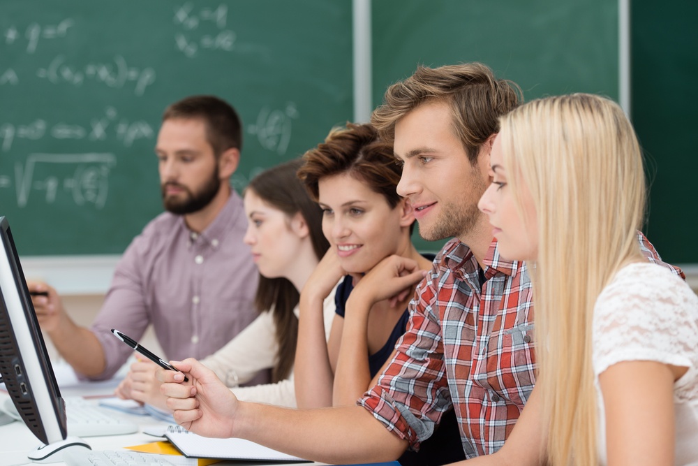 College or university students studying using a computer sitting together at a long table in the classroom reading something together on the monitor